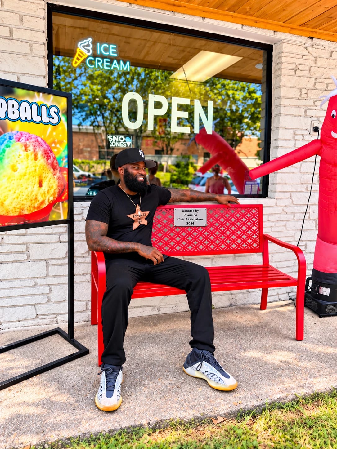 Alan Roland sits on the red bench outside POParazzi's next to a plaque reading Donated by Riverside Civic Association 2026.