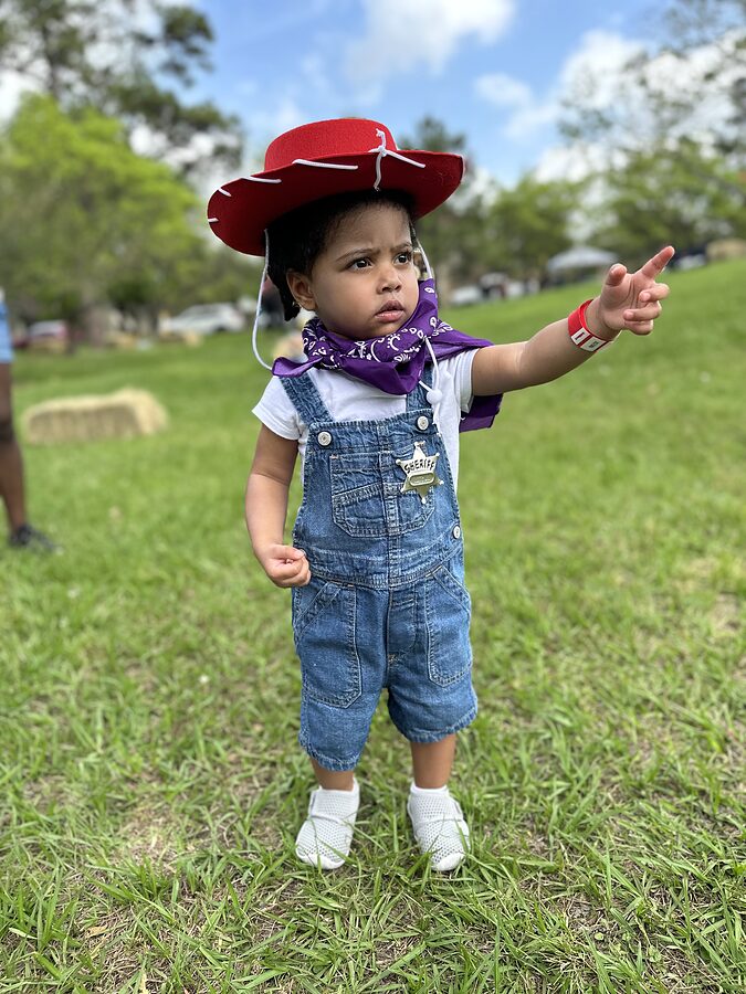 Toddler in a red cowboy hat, bandana and denim overalls points off camera on a grassy park lawn.