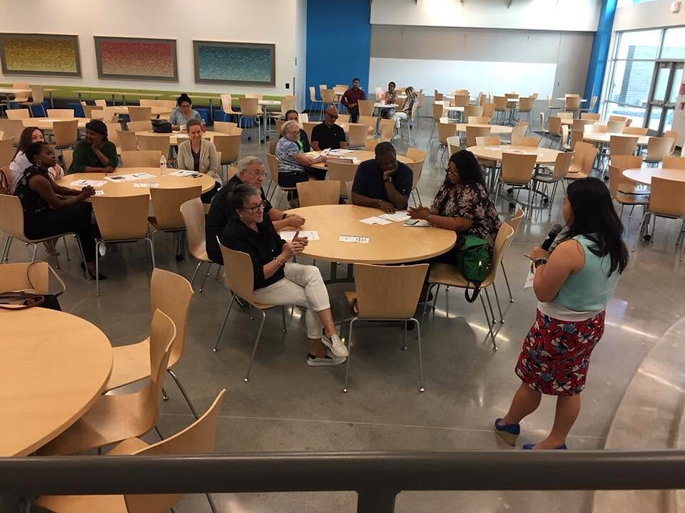 Residents seated at round tables in a school cafeteria listen to a woman speaking with a microphone.