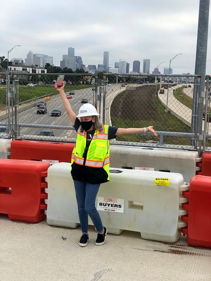 Person in hard hat and safety vest on a freeway overpass with the Houston skyline behind.