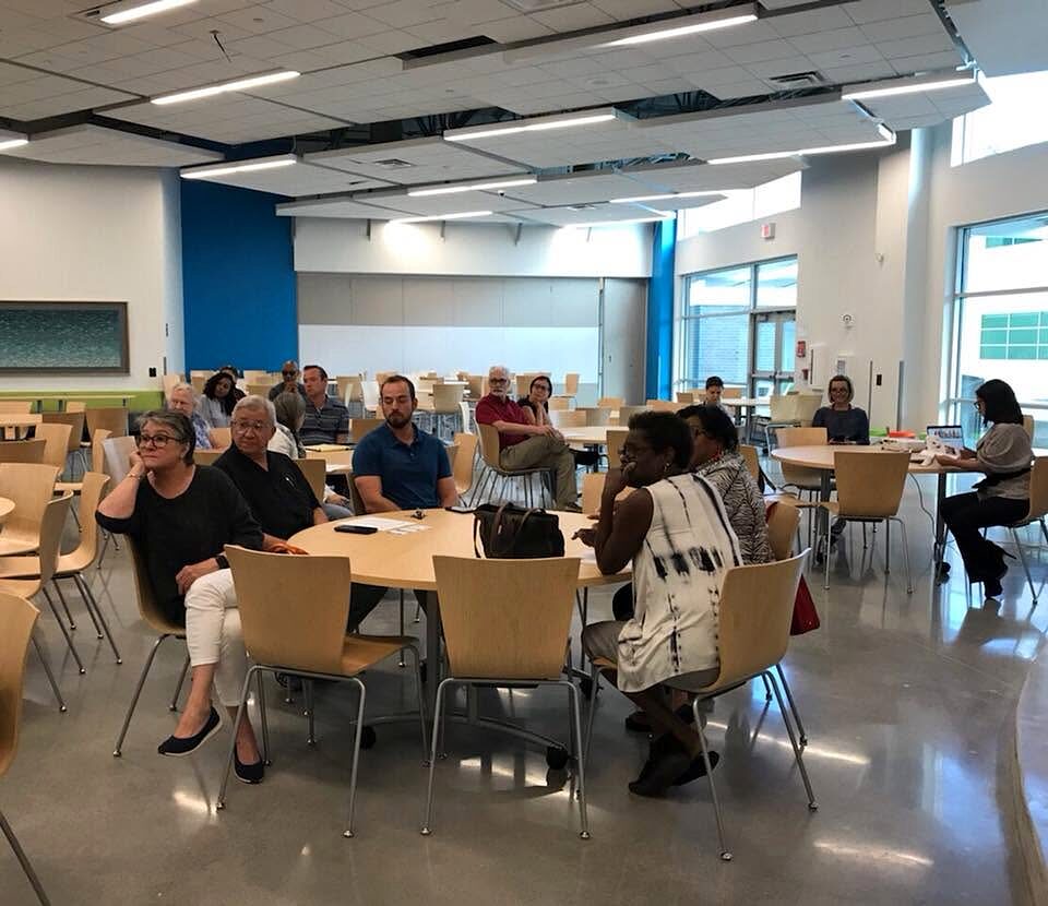 Residents seated at round tables in a bright community room listening during a neighborhood meeting.