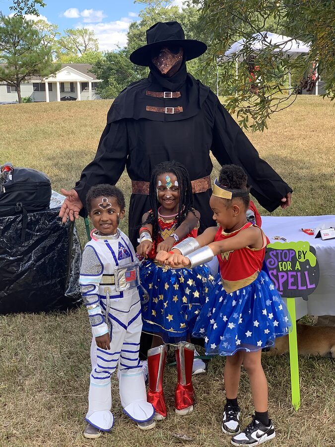 Three children in astronaut and superhero costumes pose in front of a Halloween decoration sign on a lawn.