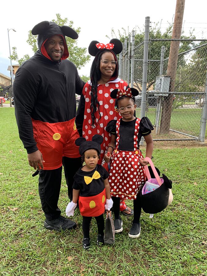 Family of four in matching mouse-themed Halloween costumes posing on grass near a chain-link fence.