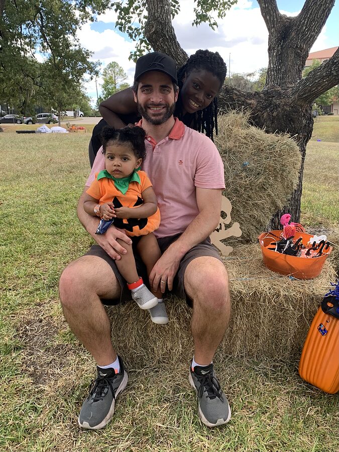 Two adults and a toddler in a pumpkin costume pose together on hay bales at an outdoor Halloween event.