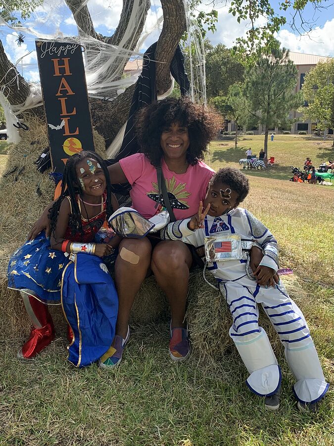 An adult and two children in Halloween costumes sit on hay bales beside a Happy Halloween sign at a park.
