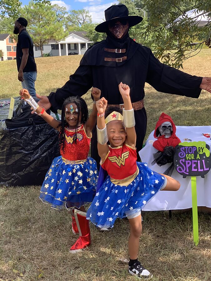 Two children in superhero costumes pose beside an adult in a cloaked costume near a decorated table.
