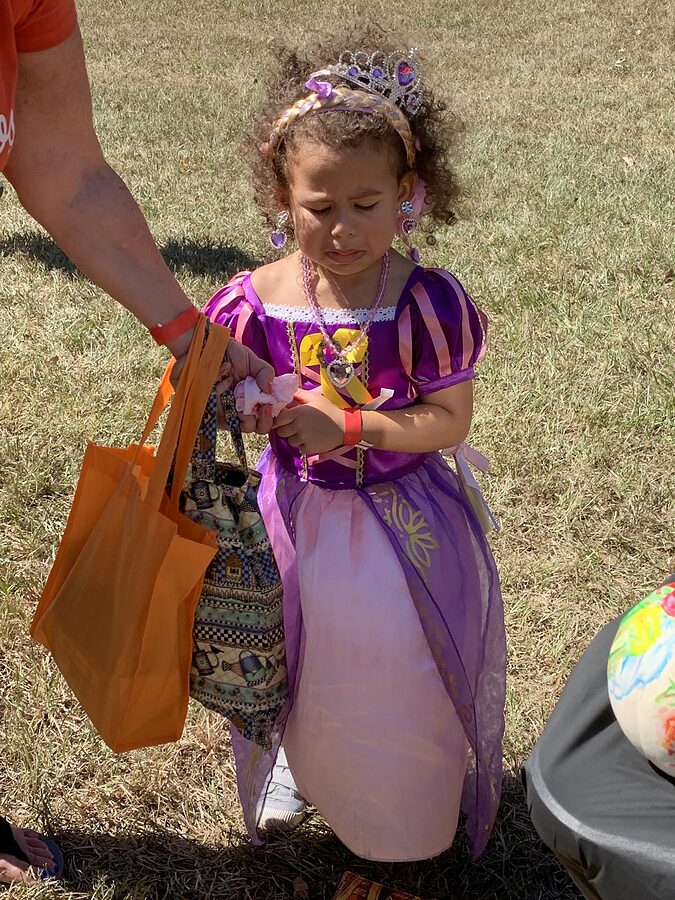Small child in a princess costume and tiara receives candy from an adult holding an orange tote bag.