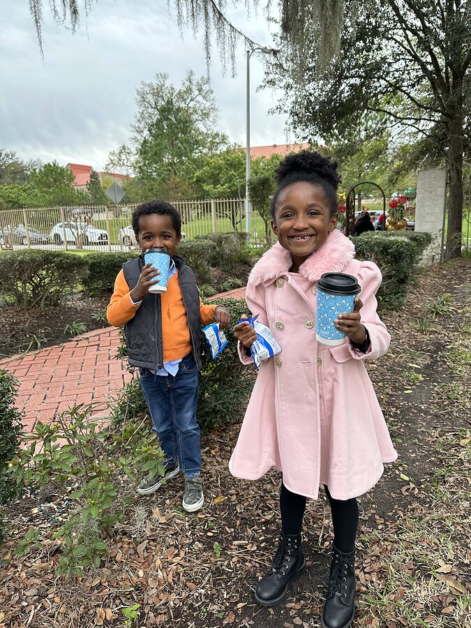 Two children in winter coats hold hot cocoa cups on a park walkway at a holiday gathering.