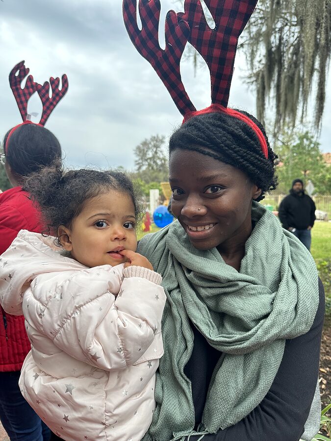 An adult wearing plaid reindeer antlers holds a toddler in a winter coat at an outdoor holiday event.