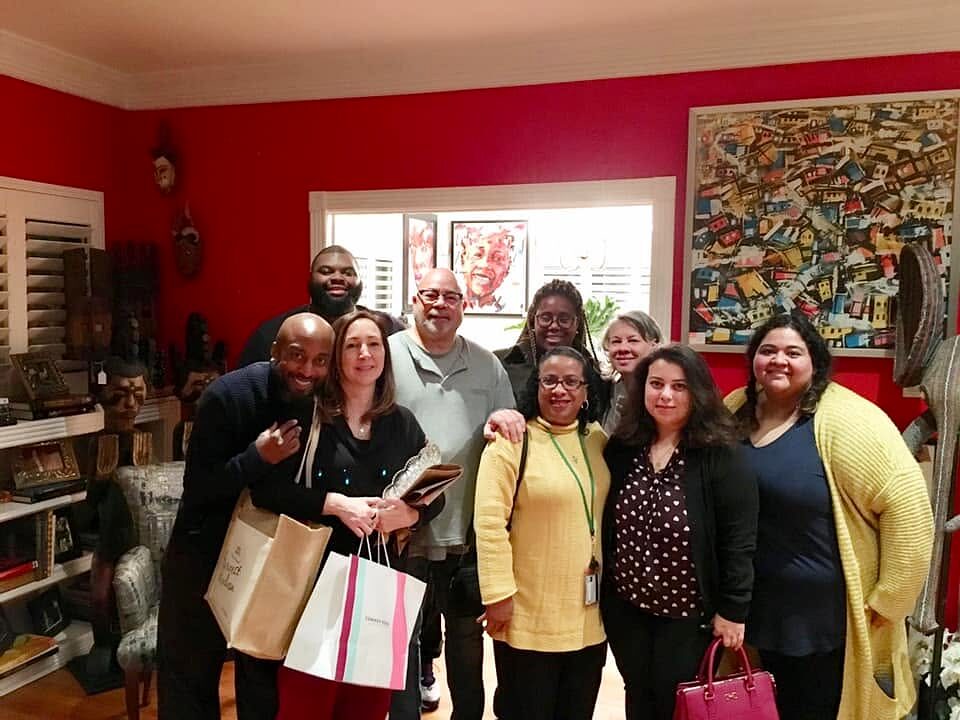 Eleven people pose indoors in a red-walled room filled with art and framed works, some holding gift bags.