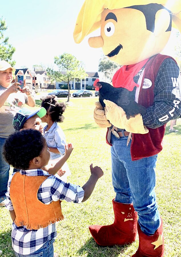 A costumed farmer mascot holds a black chicken while young children in Western vests reach toward it.