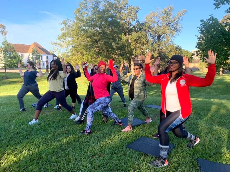 A group exercises outdoors on yoga mats in a grassy park, arms raised, under morning sun.