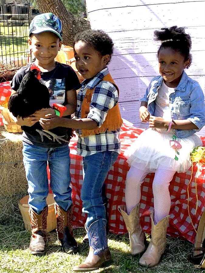 Three children in Western clothing at a red checkered table; one holds a black chicken.
