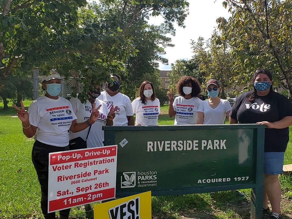 Volunteers in voter registration shirts pose behind the Riverside Park sign with an event yard sign.