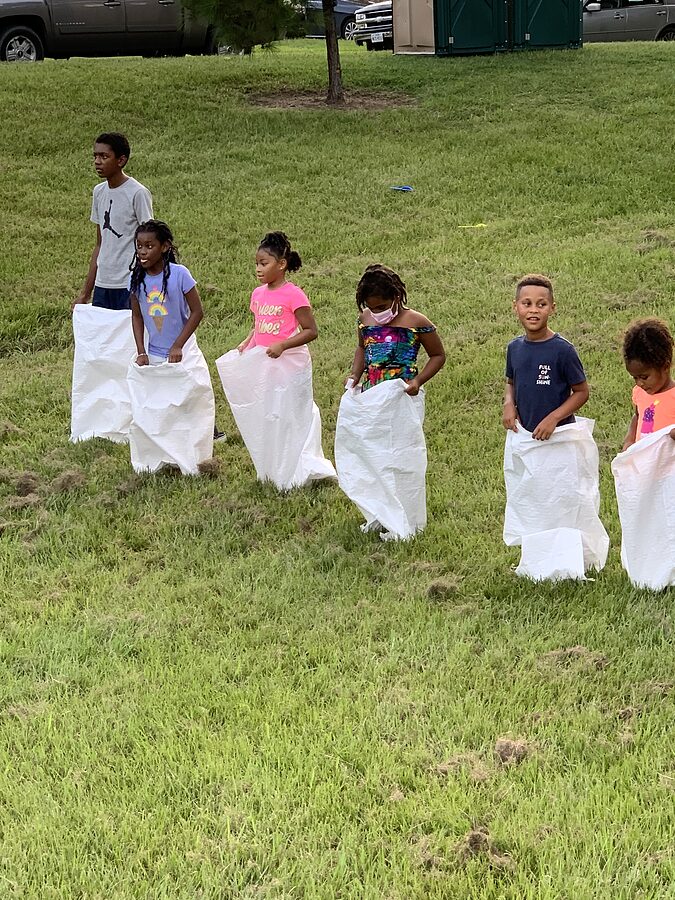 Six children holding white sacks spread across a grassy field before a race, with vehicles behind them.