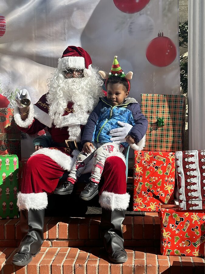 Santa Claus seated on brick steps holding a small child in an elf hat, surrounded by wrapped gifts.