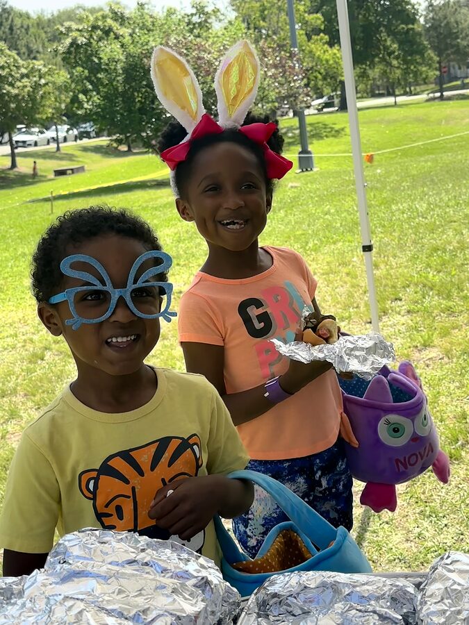 Two smiling children in bunny ears and novelty glasses holding egg-hunt baskets on a grassy park lawn.