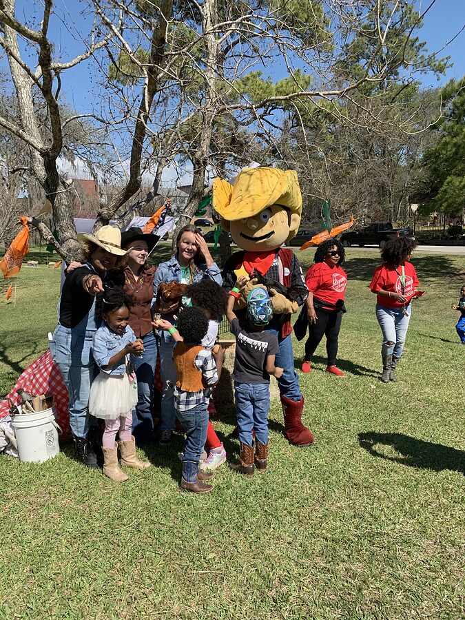 Adults in cowboy hats, children in Western outfits and a costumed mascot pose together on a park lawn.