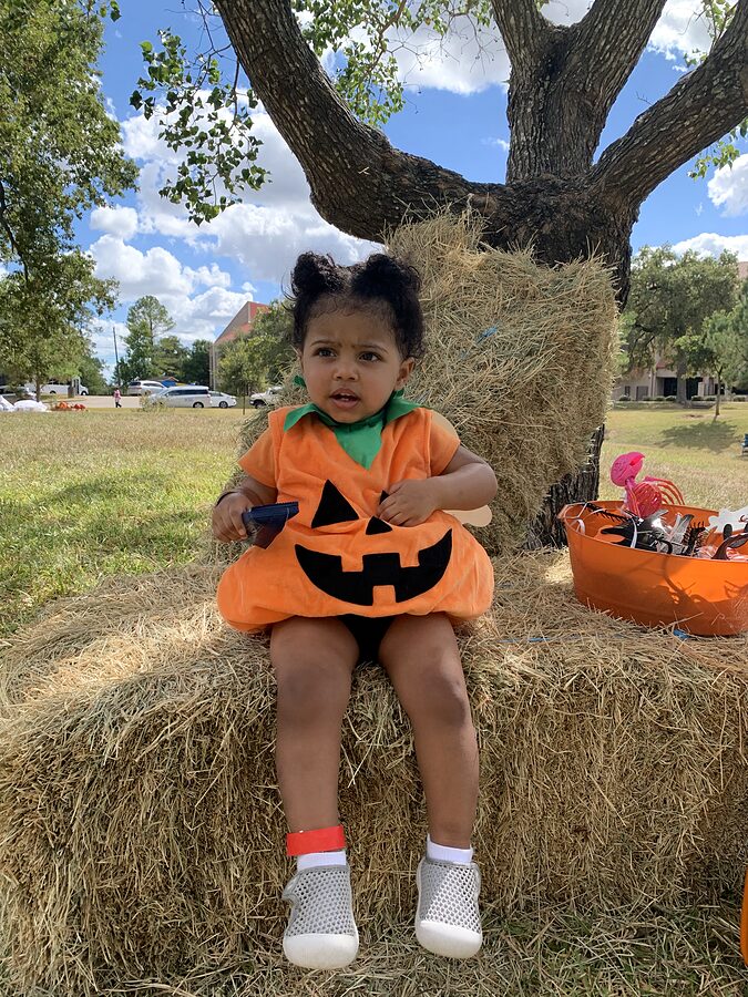 Toddler in an orange jack-o'-lantern costume sits on a hay bale under a large oak tree at a park.
