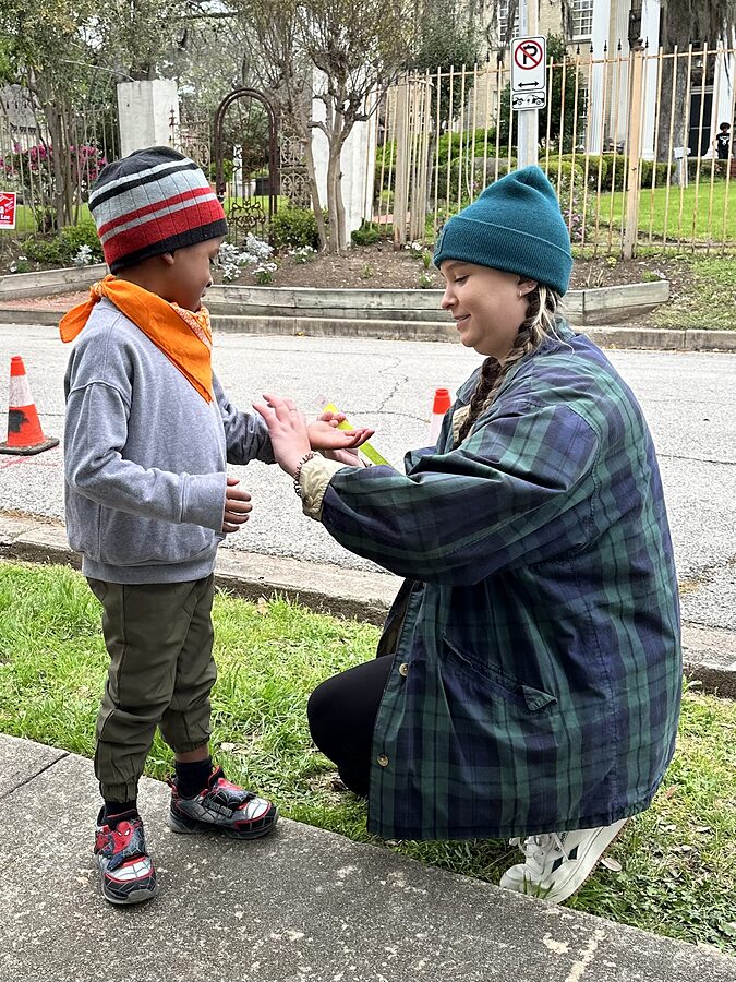 Volunteer kneeling on a curb helping a child in an orange bandana at an outdoor neighborhood event.