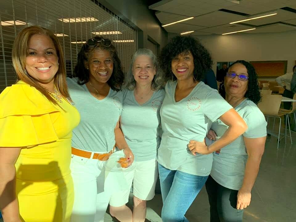 Five women in matching volunteer T-shirts and one in a yellow dress pose together indoors.