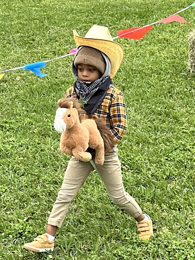 A child in a straw cowboy hat, plaid shirt and bandana carries a stuffed horse across a grass field.
