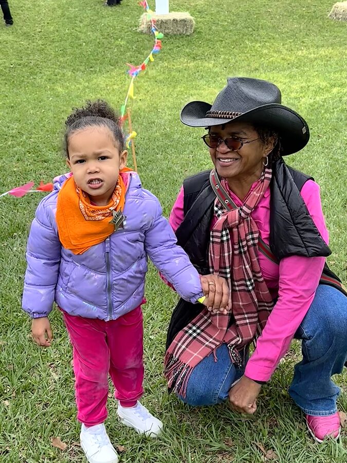 A toddler in an orange bandana stands with an adult in a cowboy hat on a grassy lawn with pennant banners.