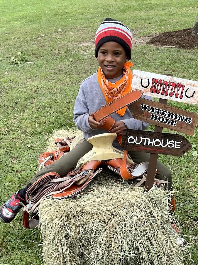 Smiling child in an orange bandana sitting on a hay bale with a leather saddle beside Western-themed signs.