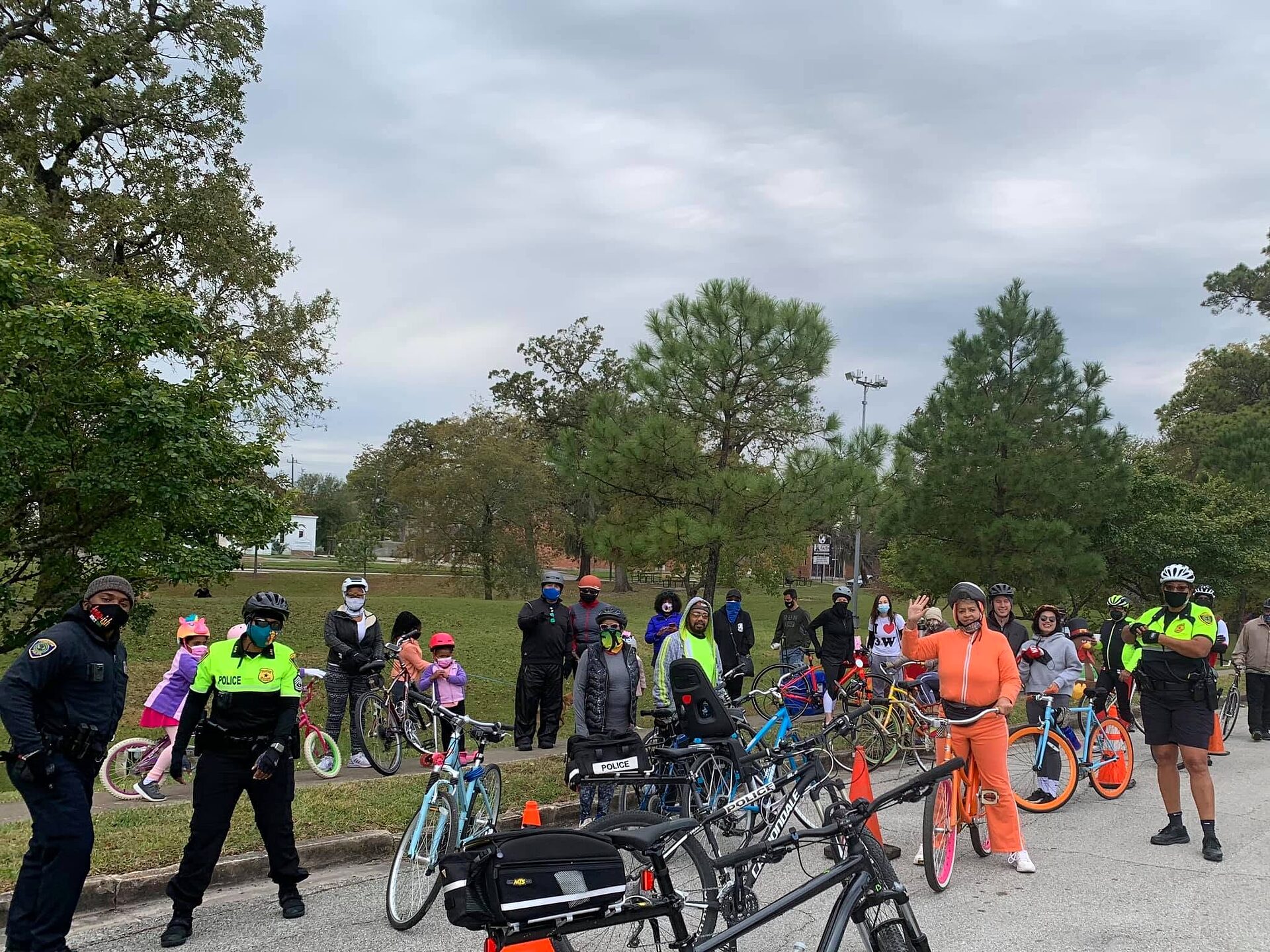 Group of residents with bicycles gathers on a park street alongside police officers and traffic cones.