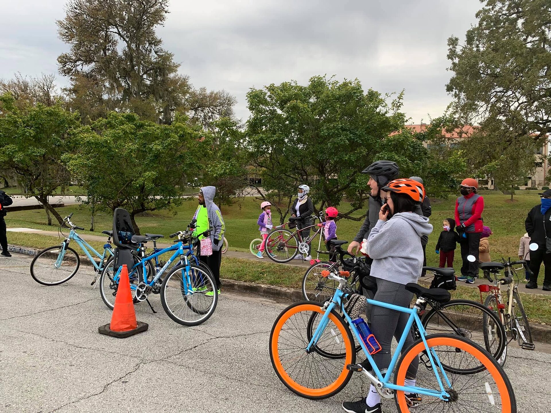 Cyclists in helmets stand with bicycles and a traffic cone on a street beside a grassy park.