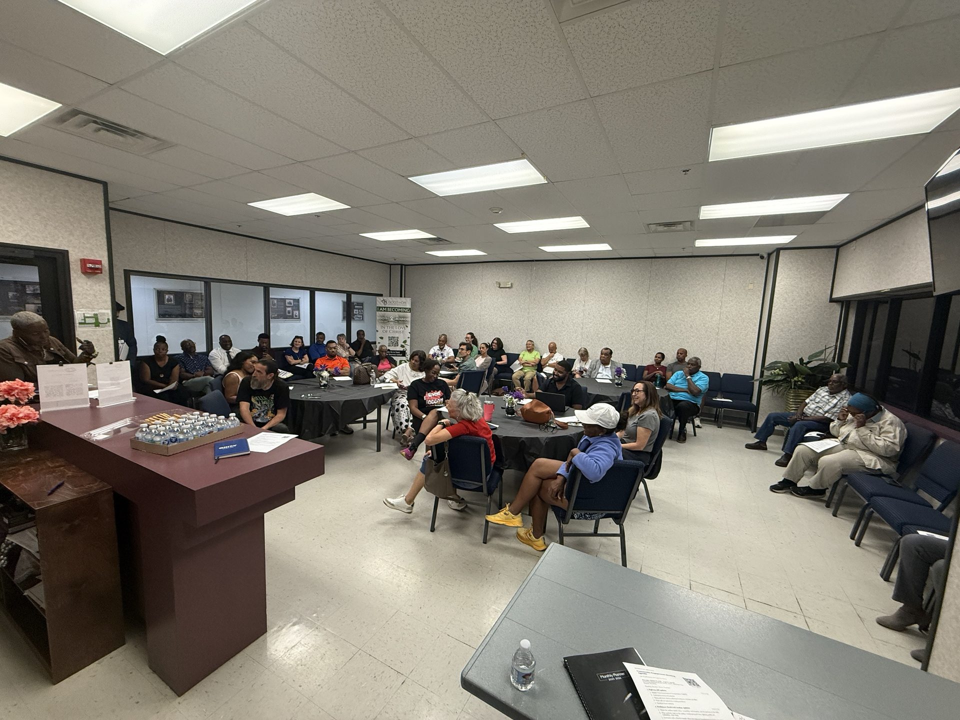 Neighbors seated at round tables with black tablecloths and purple flower arrangements in the Good Hope Church fellowship hall, listening to a speaker at the podium during the RCA Community Engagement Meeting on October 8, 2026.
