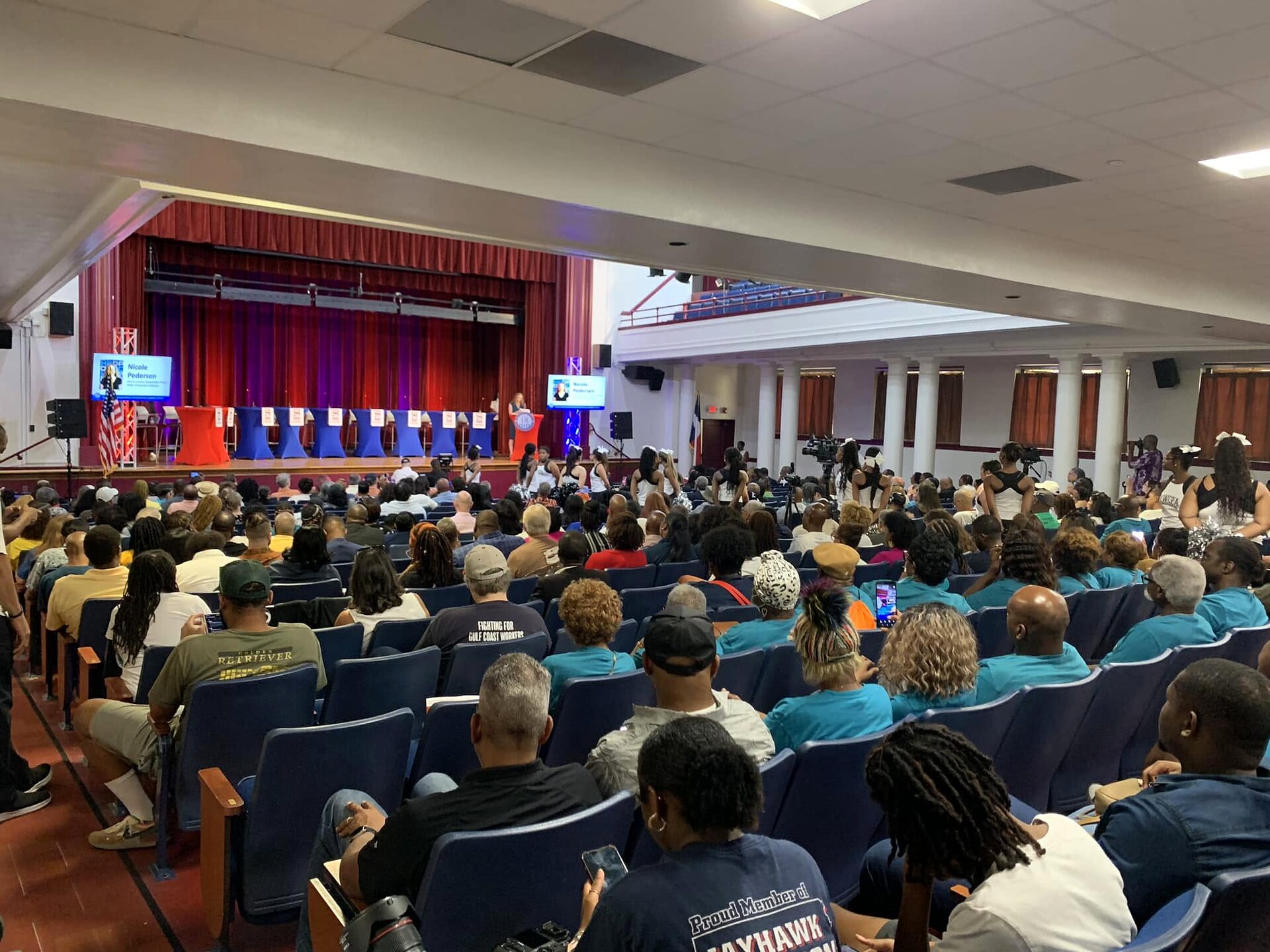 Large audience filling a school auditorium facing a stage with seated panelists and a screen.