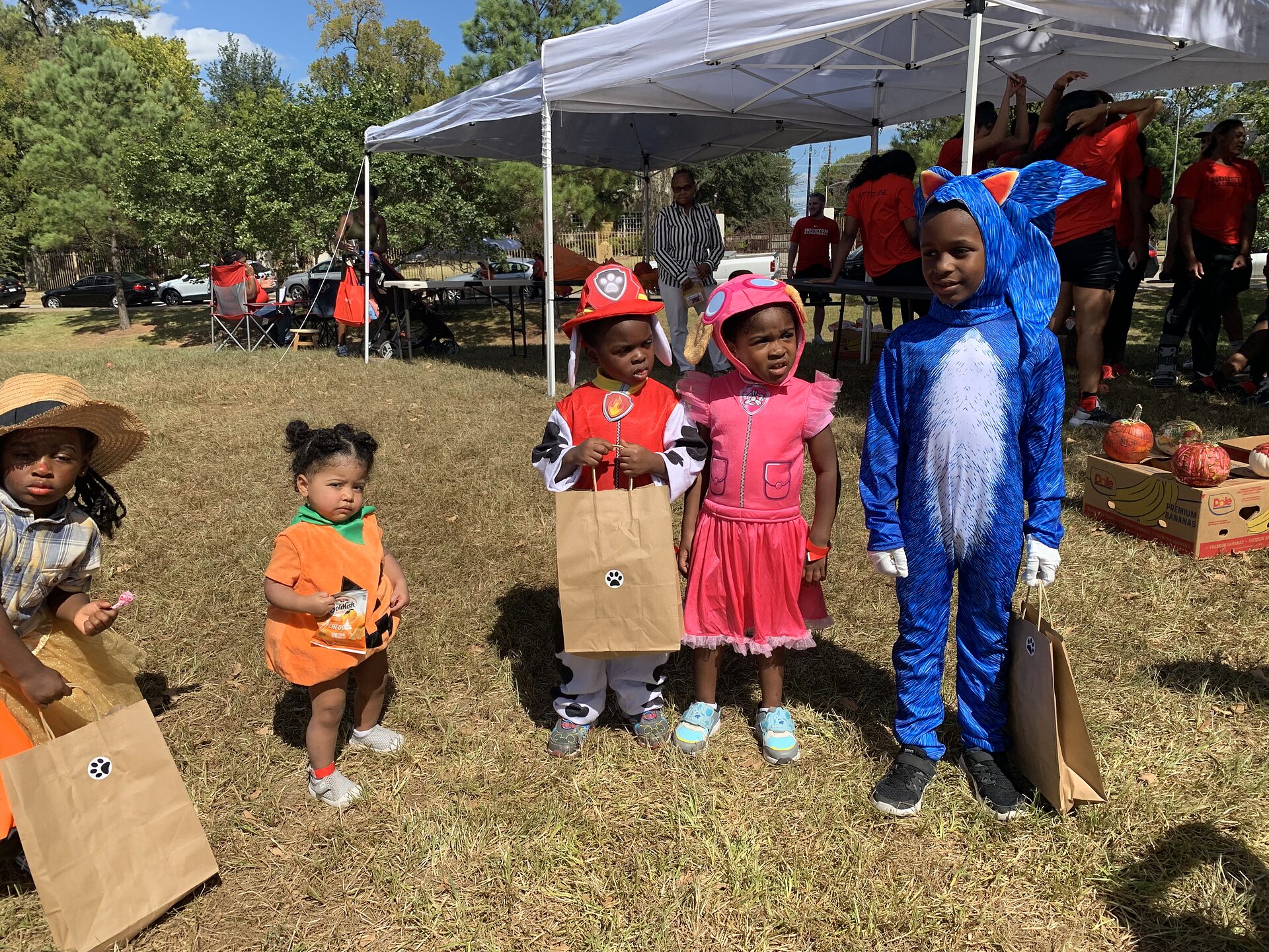 Five children in Halloween costumes stand on a park lawn holding treat bags, with a canopy and pumpkins behind.