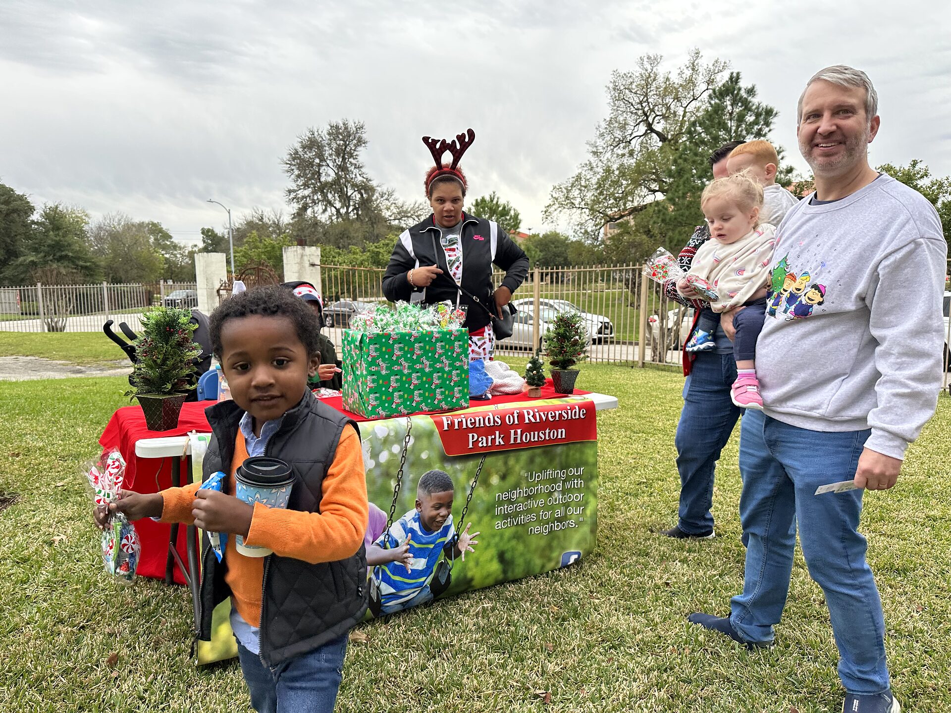 Volunteers and families at an outdoor holiday table with wrapped gifts and a Friends of Riverside Park banner.