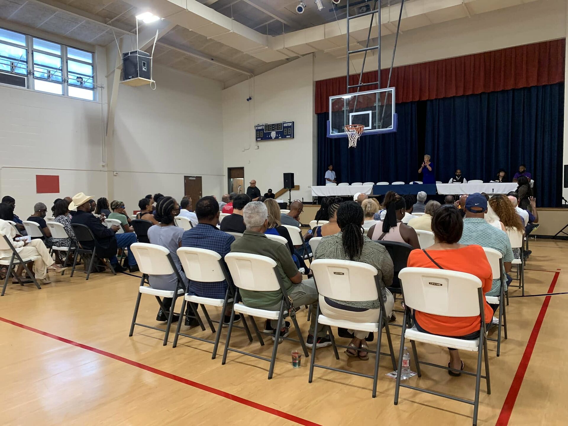 Residents seated in folding chairs in a gymnasium facing a head table and a speaker.