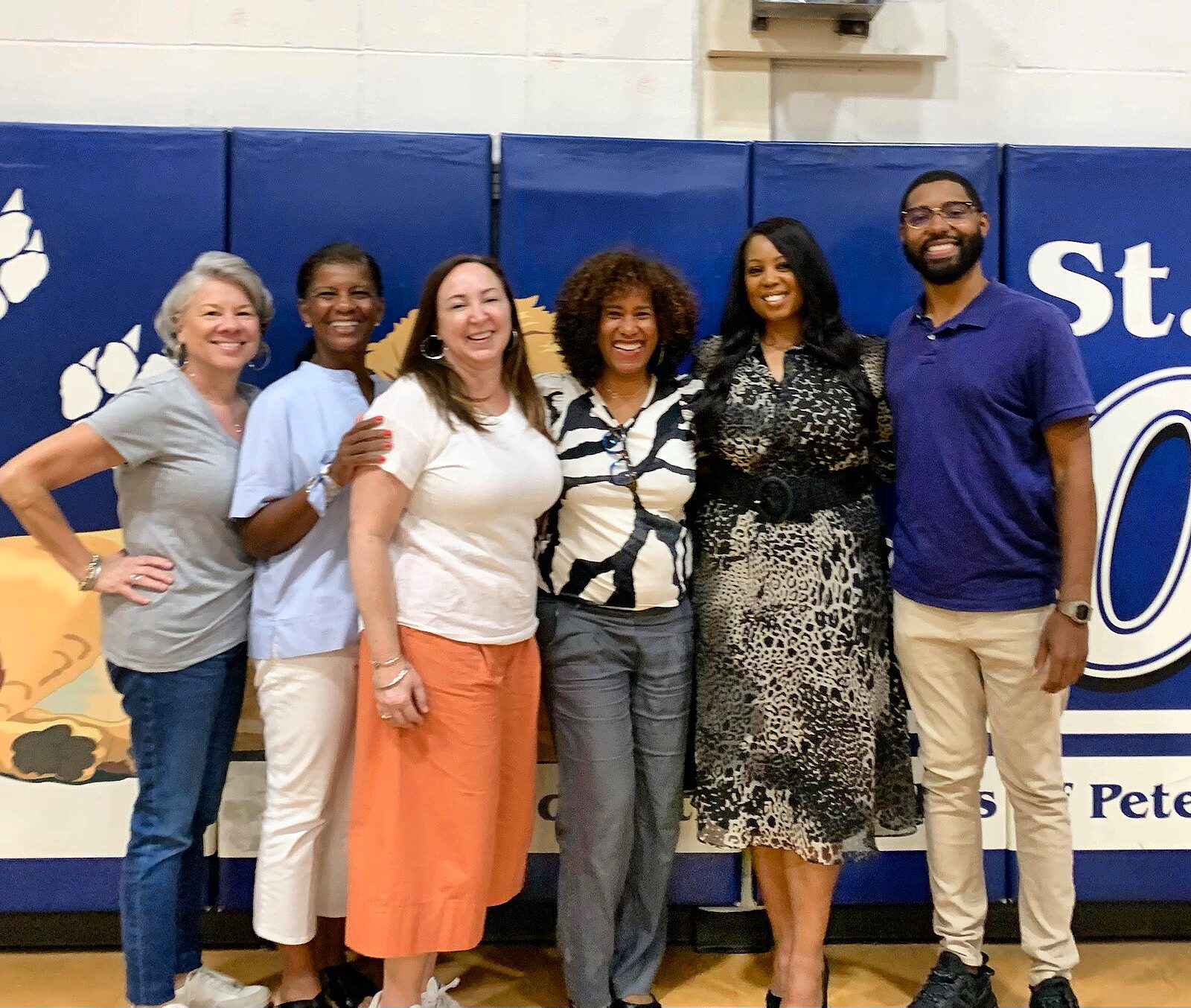 Six adults pose together in a school gymnasium in front of blue wall padding.