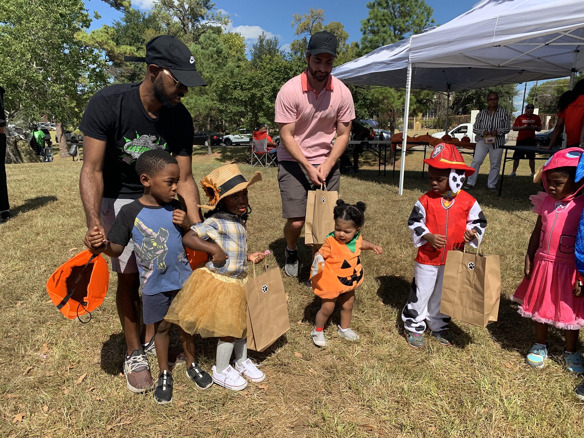 Adults and costumed children with treat bags walk across a park lawn near a white event canopy.