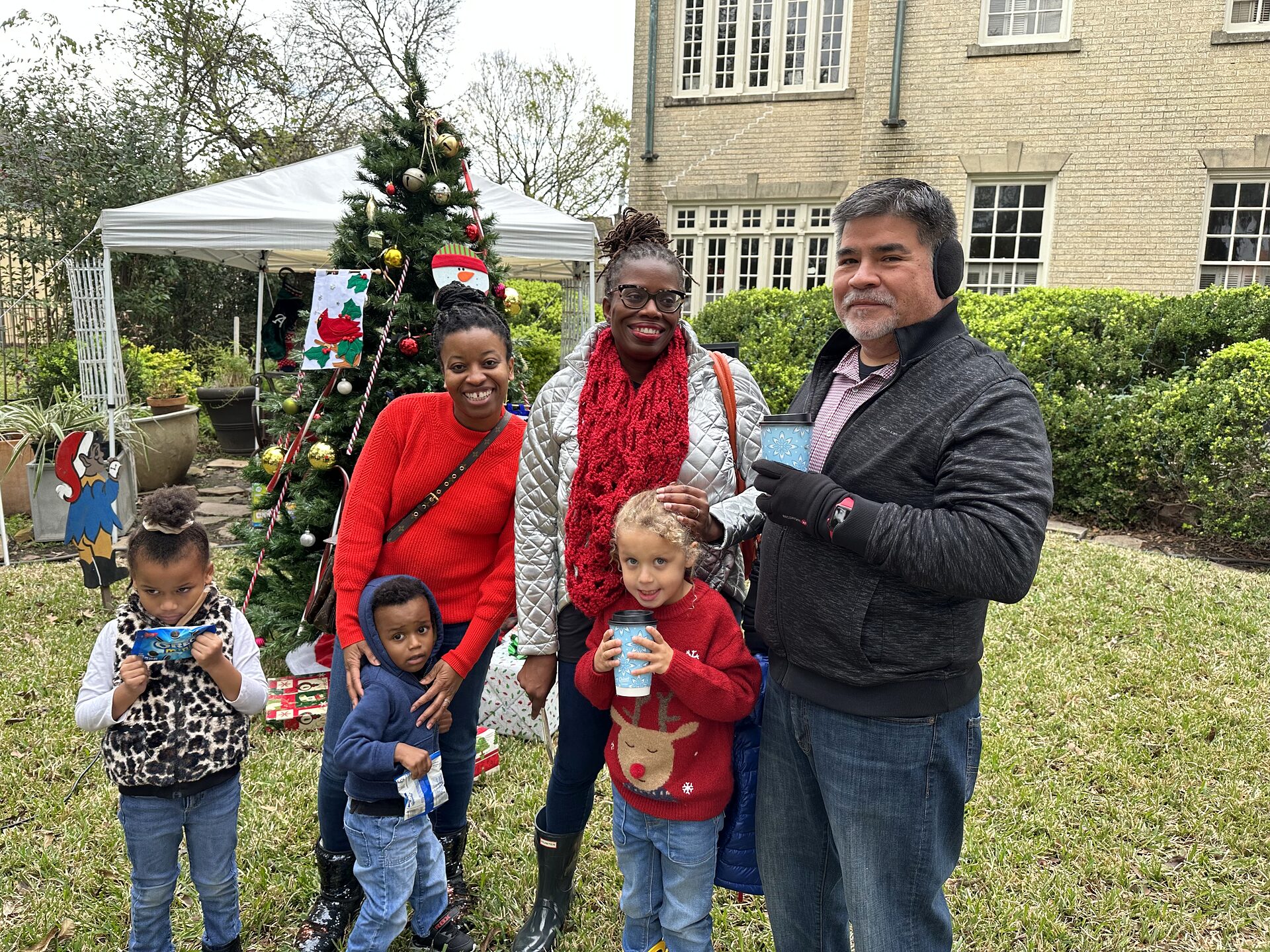 Families with young children pose beside a decorated Christmas tree and white tent on a lawn.