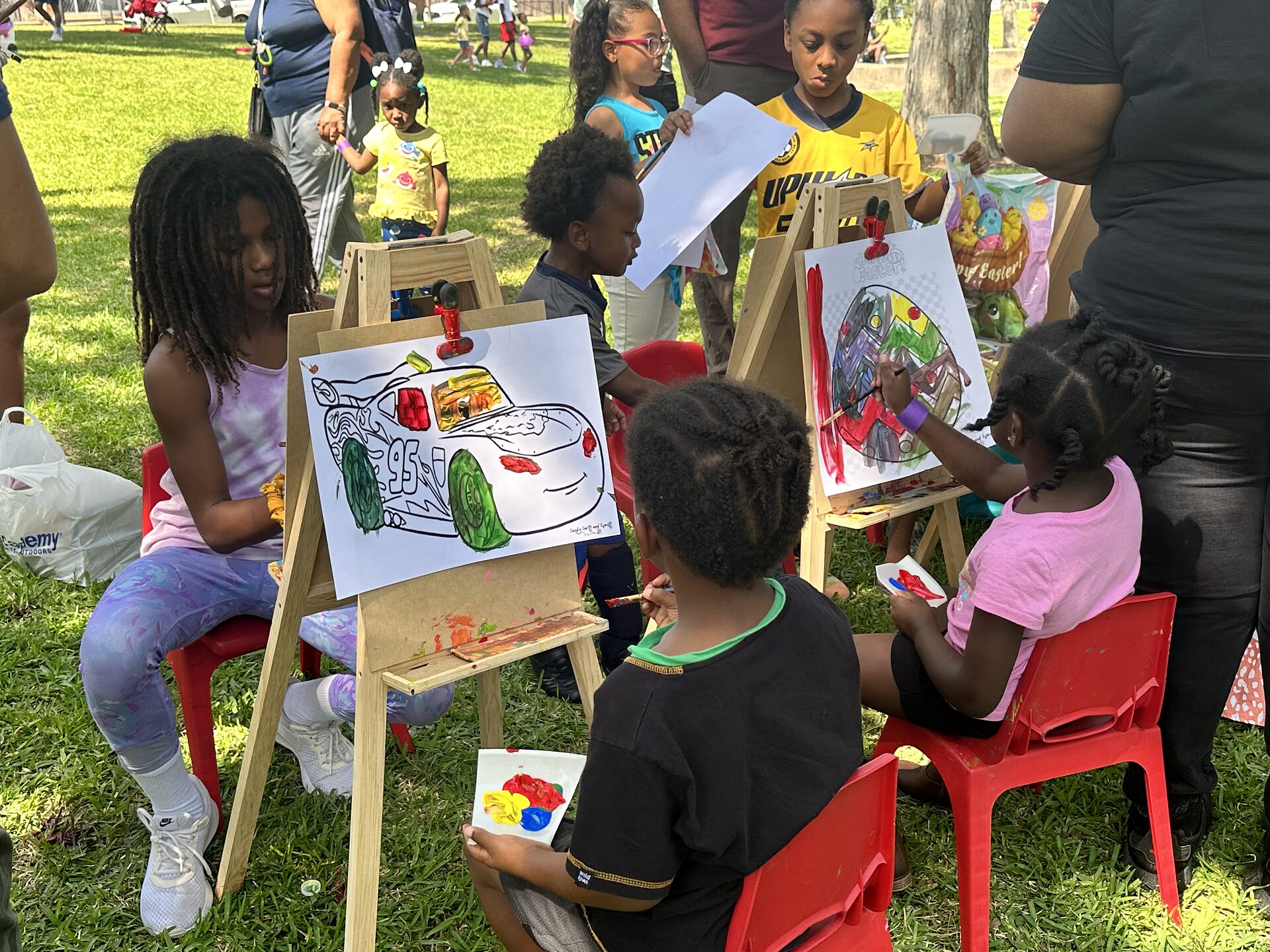 Children painting pictures on wooden easels with red chairs at an outdoor park event.