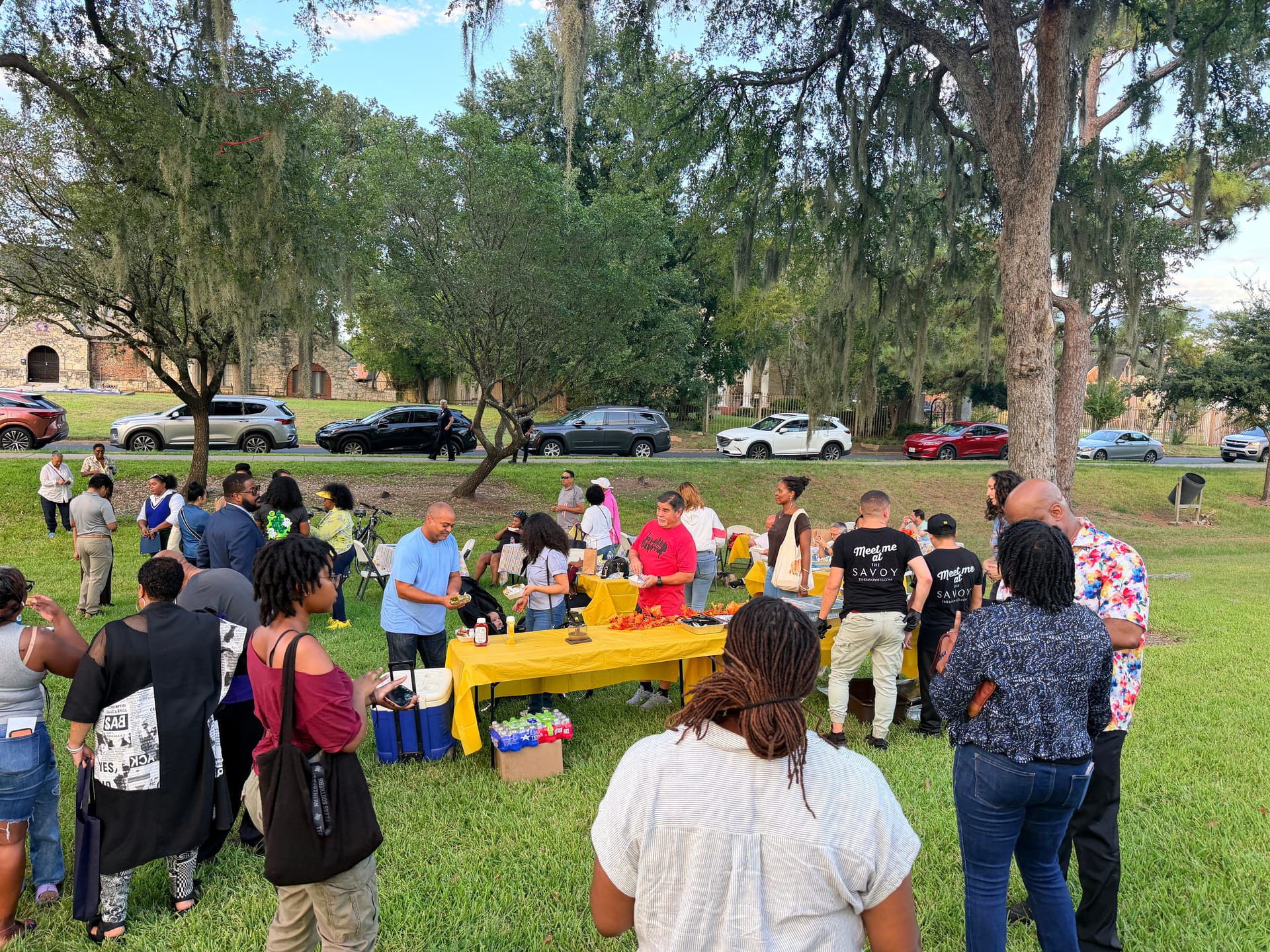 Neighbors gather around food tables under the oak trees at Riverside Park for National Night Out, October 6, 2026.