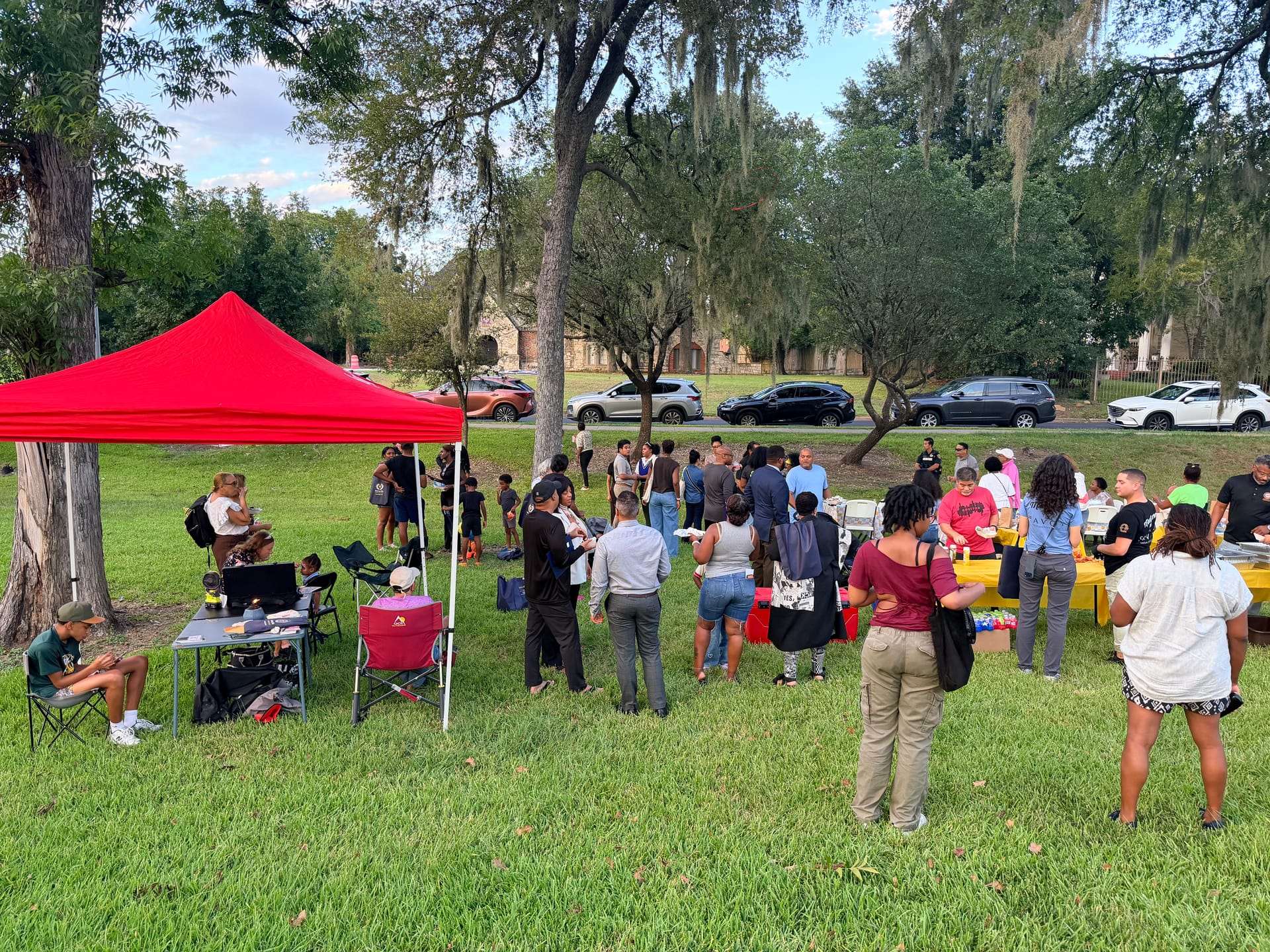 A crowd of neighbors and families gathers on the lawn at Riverside Park around a red canopy and yellow-covered food tables under oak trees.