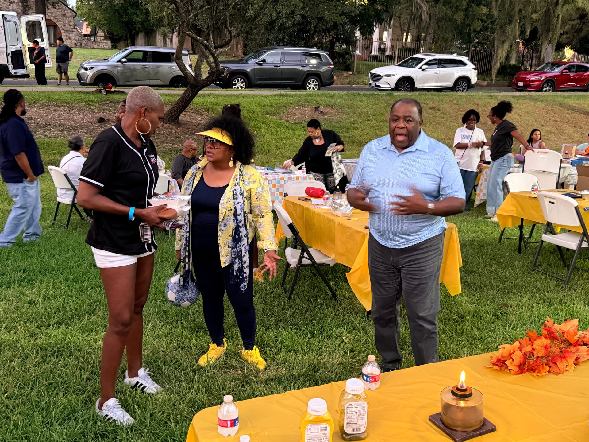 State Representative Jolanda Jones, Tomoro Bell in a yellow jacket, and Council Member Willie Davis, At-Large 2, in a light blue shirt, talk at dusk beside a yellow-covered table with a lit candle at Riverside Park.