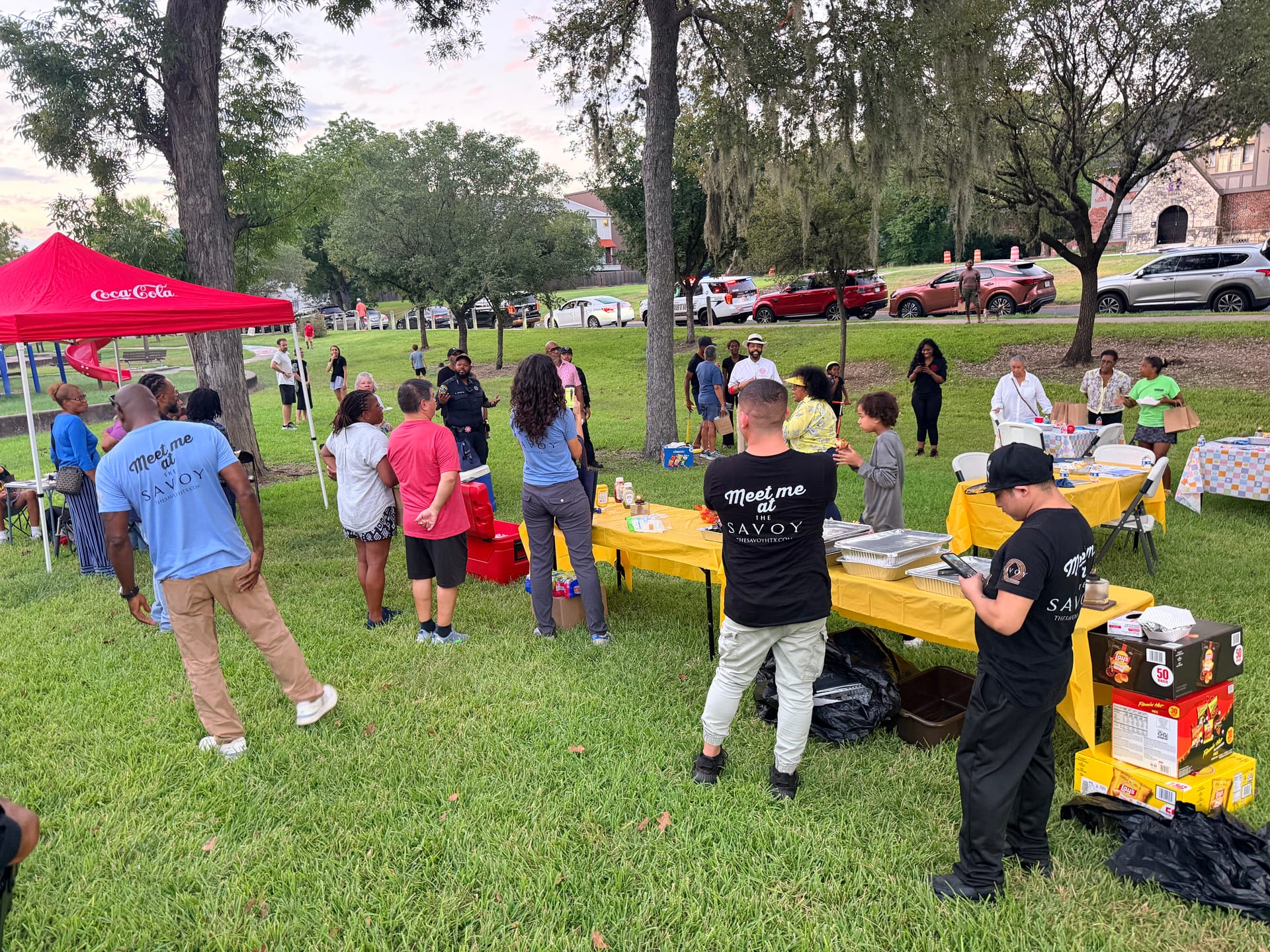 Neighbors gather around yellow-covered food tables under oak trees at Riverside Park, with a red Coca-Cola canopy at left and volunteers in Meet me at The Savoy shirts serving food.