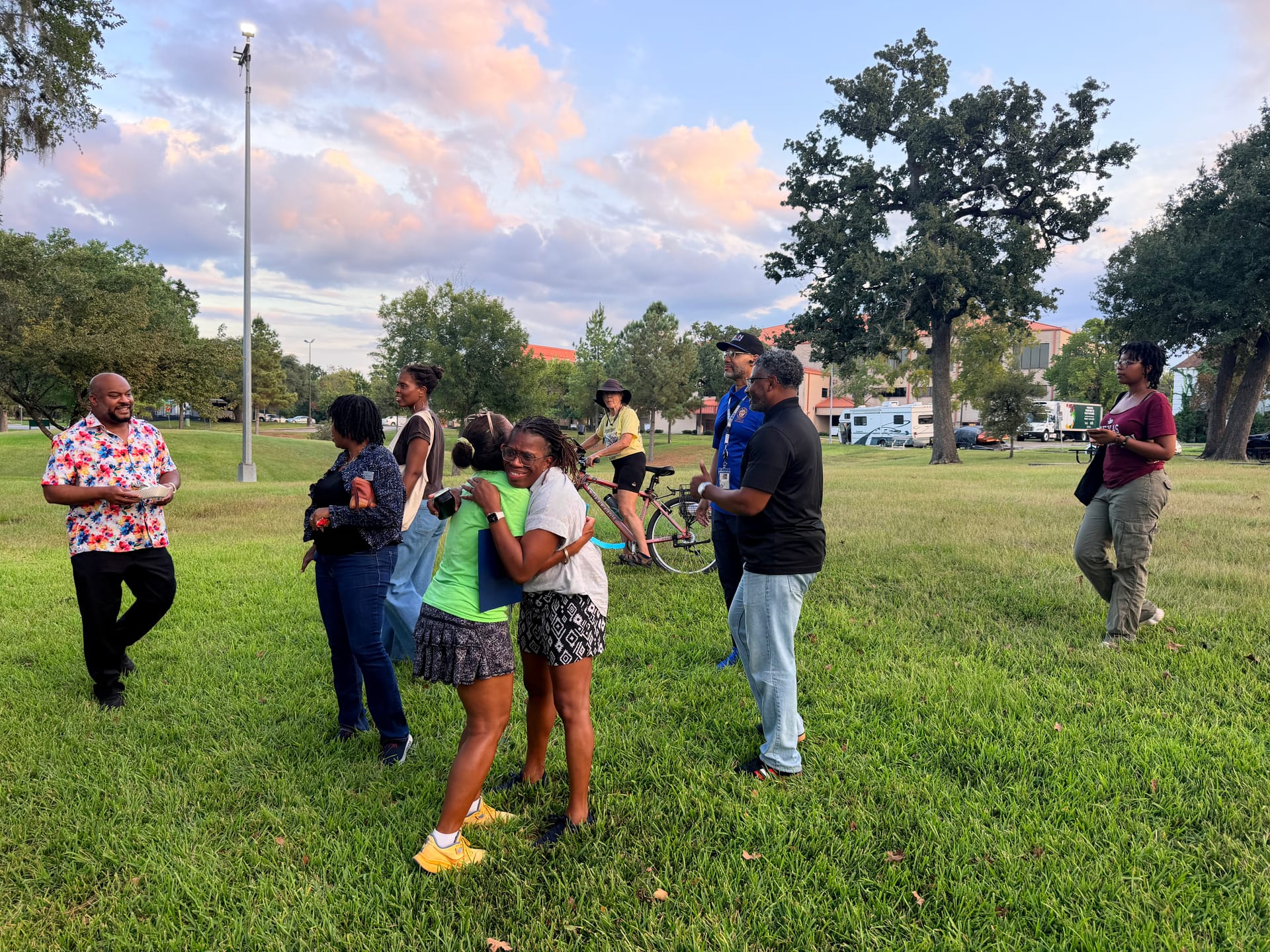 RCA Vice President Sharon Evans-Brooks, in a neon green shirt, hugs a neighbor on the lawn at Riverside Park under a pink sunset sky as other neighbors arrive.