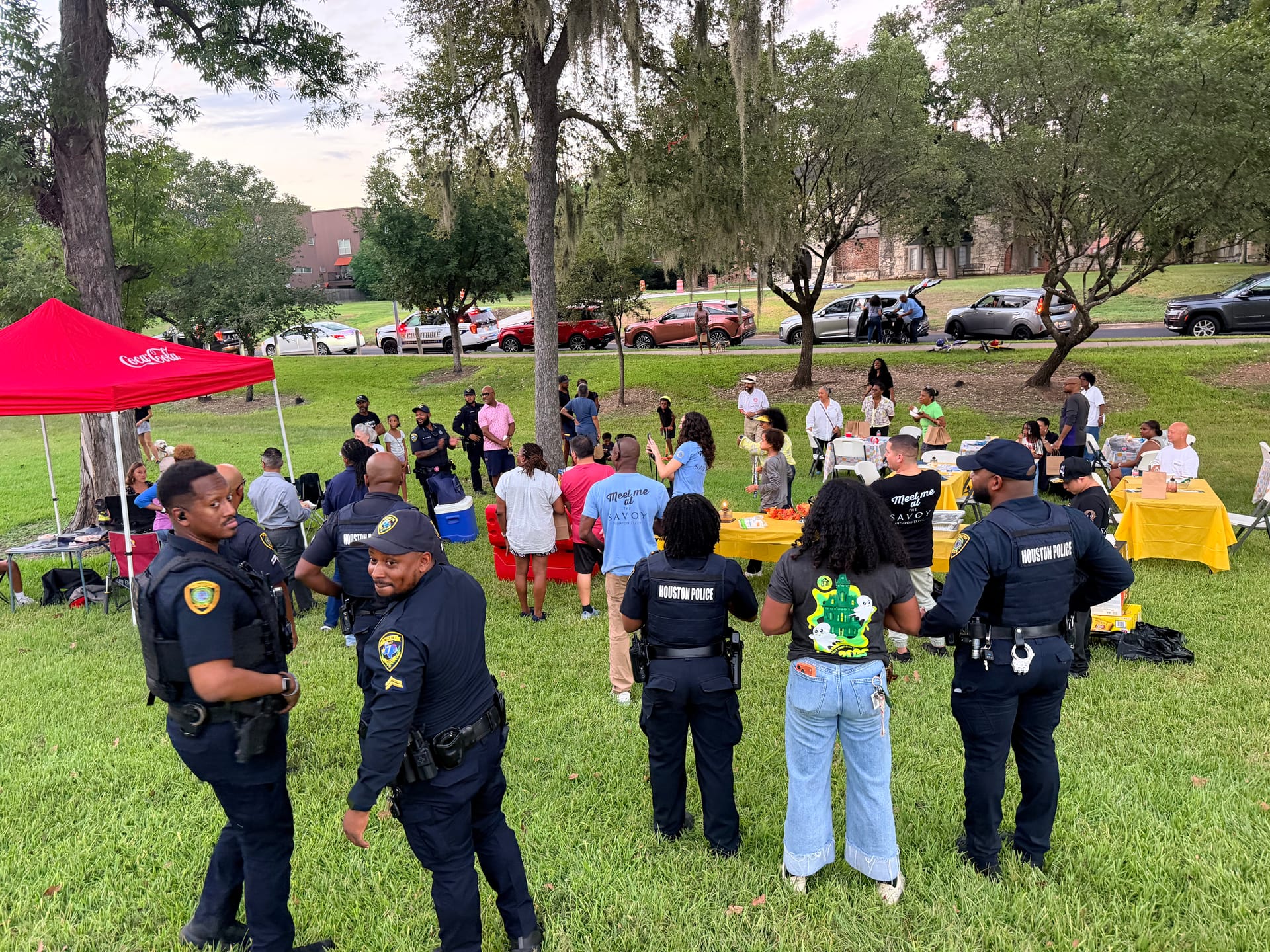 Uniformed Houston Police officers stand on the lawn at Riverside Park as neighbors gather around the red canopy and yellow-covered food tables.