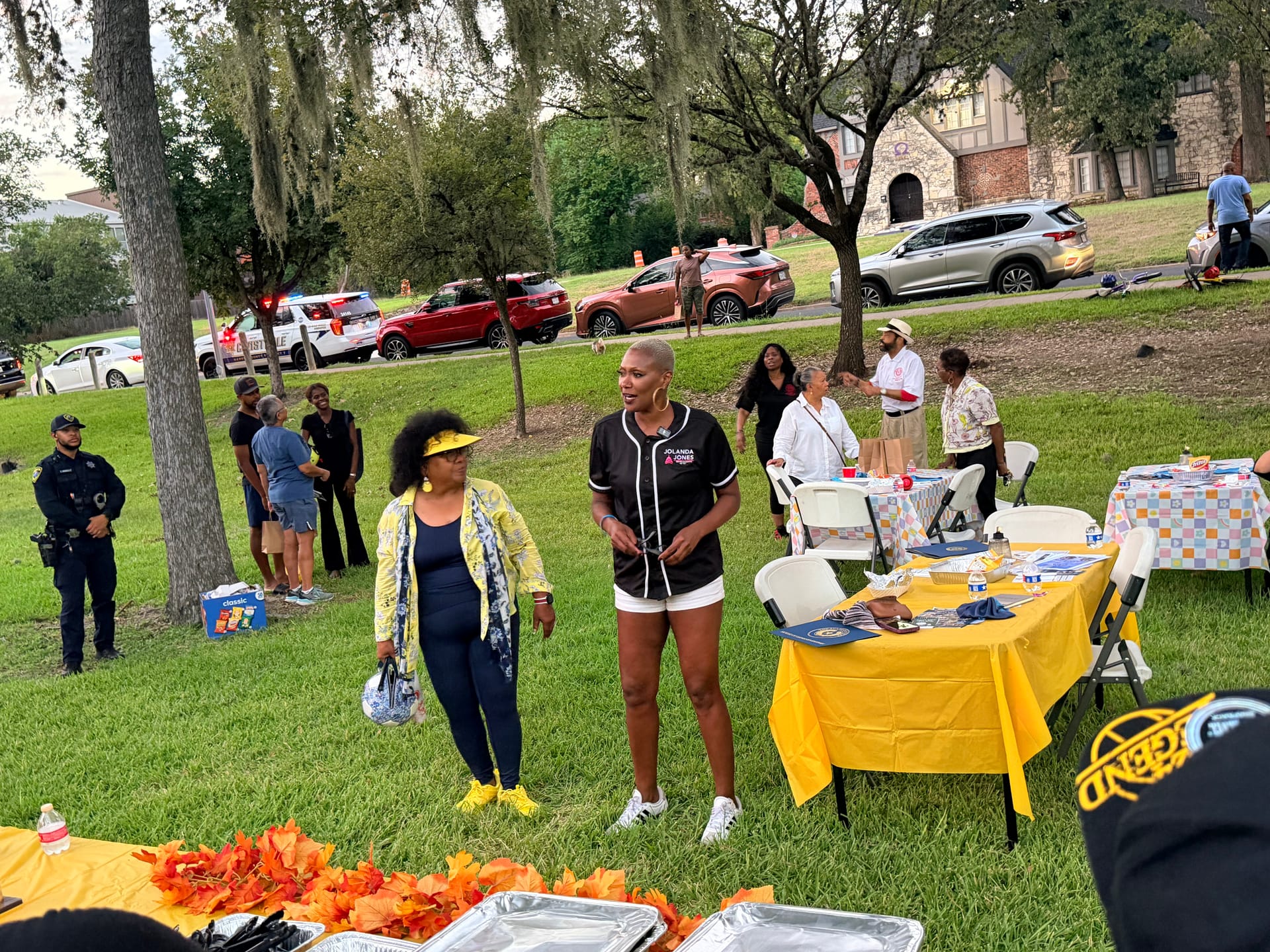 State Representative Jolanda Jones, in a black jersey, talks with Tomoro Bell, in a yellow jacket, beside yellow-covered tables at Riverside Park while an HPD officer stands nearby.