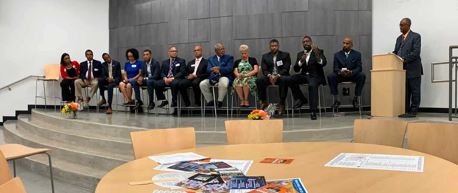 A row of seated panelists in business attire faces the room while a man speaks at a podium.