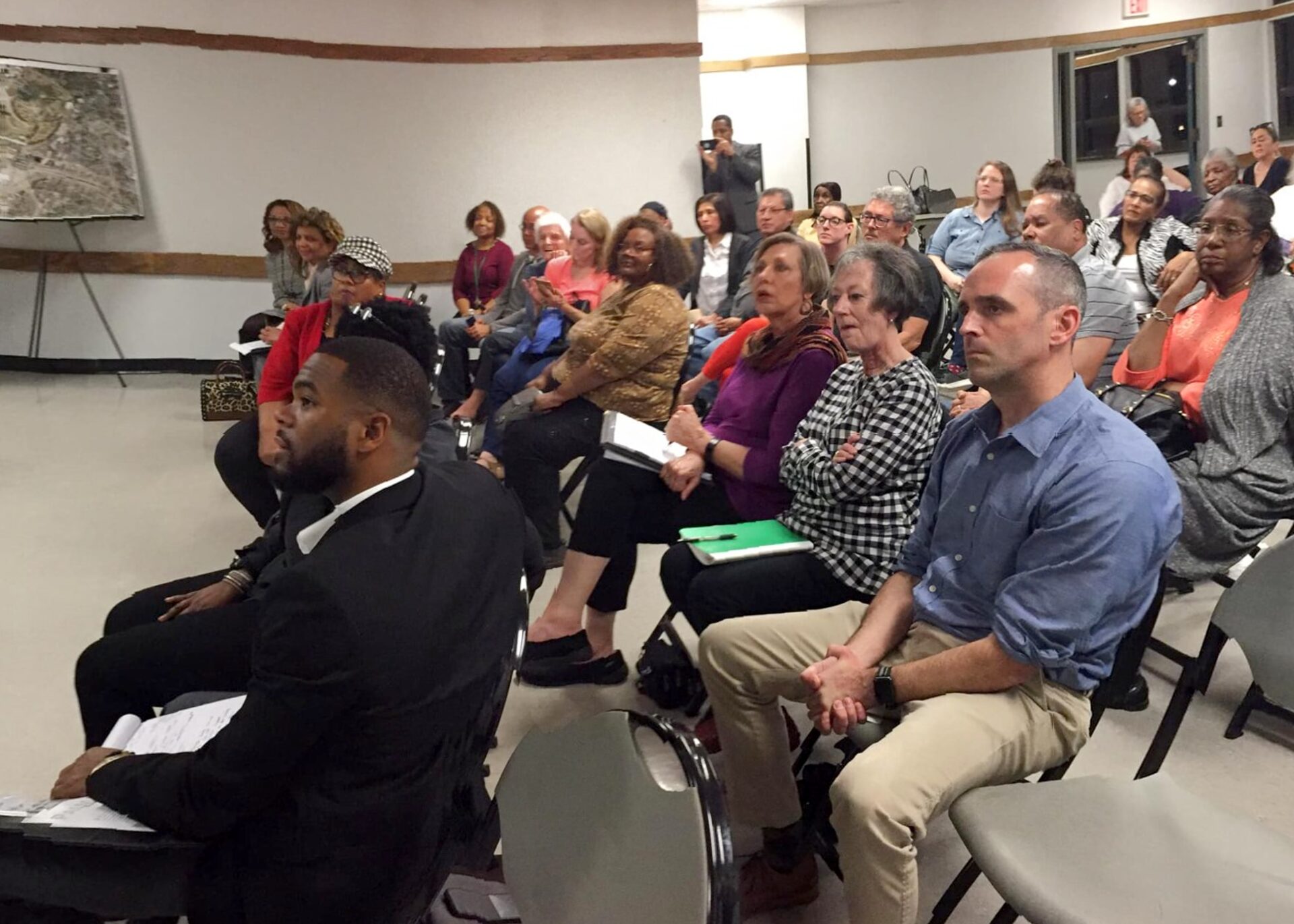 Rows of seated residents listening attentively at an indoor community meeting with a map on the wall.