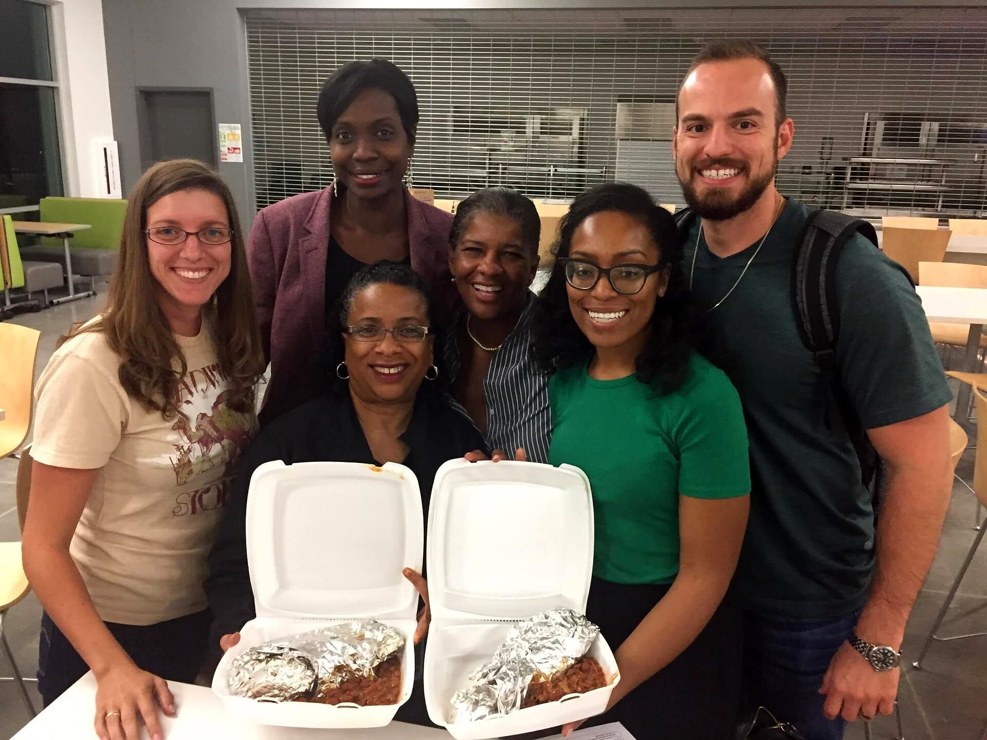 Six volunteers pose together indoors behind two open takeout containers of food on a table.