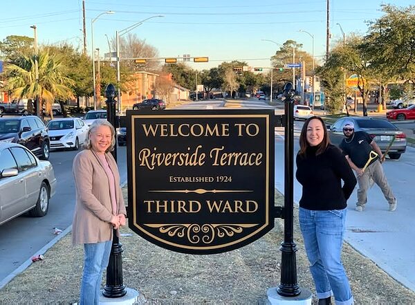 Two women stand beside the black and gold Welcome to Riverside Terrace neighborhood sign on a median.